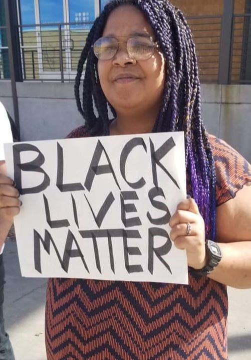 Person with long black and purple braids, glasses, brown skin, orange and black patterned shirt, holding a sign that reads Black Lives Matter.