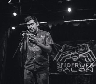 Person standing on stage in front of backdrop that reads Spiderweb Salon. Person is reading into a microphone and has dark short hair and light brown skin. Person is wearing a button up shirt and pants.
